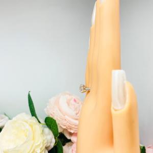 Mannequin hand with long white shimmery nails, displayed against a soft white-gray background with pink and white floral accents. The hand is wearing a near colorless (G/H color) round brilliant cut cubic zirconia solitaire ring in a split prong cathedral setting. The ring is made of sterling silver and features a sleek, minimalist design. The setting is simple yet elegant, allowing the center stone to take center stage.