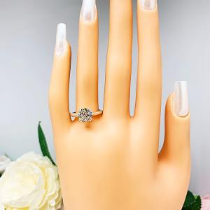 Mannequin hand with long white shimmery nails, displayed against a soft white-gray background with pink and white floral accents. The hand is wearing a near colorless (G/H color) round brilliant cut cubic zirconia solitaire ring in a split prong cathedral setting. The ring is made of sterling silver and features a sleek, minimalist design. The setting is simple yet elegant, allowing the center stone to take center stage.