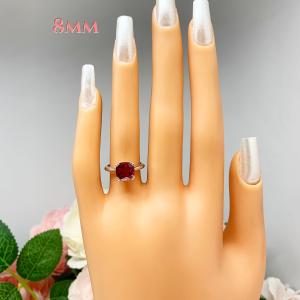 Mannequin hand with long white shimmery nails, displayed against a soft white-gray background with pink and white floral accents. The hand is wearing a sterling silver ring with a cushion cut lab created deep red ruby set in a split-prong setting. The text '8MM' appears above the hand.