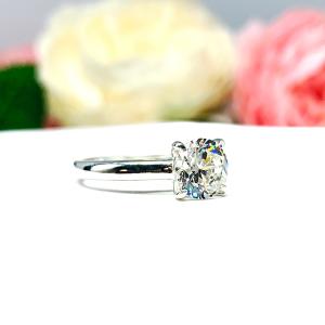 Close-up view of a near colorless (G/H color) round brilliant cut cubic zirconia solitaire ring in a claw prong cathedral setting. The ring is made of sterling silver and features a sleek, minimalist design. The setting is simple yet elegant, allowing the center stone to take center stage. The ring is displayed against a soft, blurred background of pastel-colored flowers, creating a romantic and delicate ambiance.