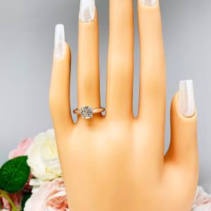 Mannequin hand with long white shimmery nails, displayed against a soft white-gray background with pink and white floral accents. The hand is wearing a near colorless (G/H color) round brilliant cut cubic zirconia solitaire ring in a claw prong cathedral setting. The ring is made of sterling silver and features a sleek, minimalist design. The setting is simple yet elegant, allowing the center stone to take center stage.