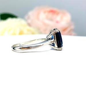 Sterling silver ring featuring a vivid elongated cushion-cut lab created blue sapphire held in a split-prong setting, with soft-focus pink and white flowers in the background.