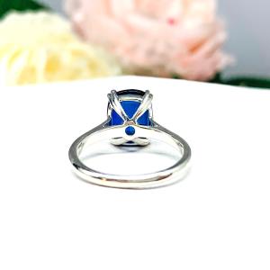 Sterling silver ring featuring a vivid elongated cushion-cut lab created blue sapphire held in a split-prong setting, with soft-focus pink and white flowers in the background.