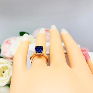 Mannequin hand with long white shimmery nails, displayed against a soft white-gray background with pink and white floral accents. The hand is wearing a sterling silver ring featuring a vivid elongated cushion-cut lab created blue sapphire held in a split-prong setting.