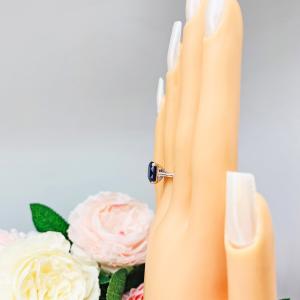 Mannequin hand with long white shimmery nails, displayed against a soft white-gray background with pink and white floral accents. The hand is wearing a sterling silver ring featuring a vivid elongated cushion-cut lab created blue sapphire held in a split-prong setting.