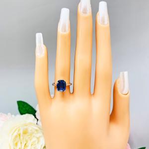 Mannequin hand with long white shimmery nails, displayed against a soft white-gray background with pink and white floral accents. The hand is wearing a sterling silver ring featuring a vivid elongated cushion-cut lab created blue sapphire held in a split-prong setting.