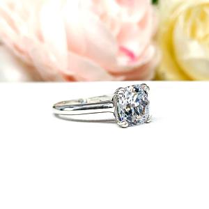 A sterling silver split-prong cathedral engagement ring set with a colorless old mine cut cubic zirconia. The ring is set against a white background with a blurred image of pink and white flowers in the background.