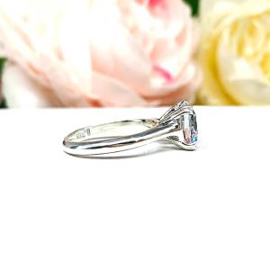 A sterling silver split-prong cathedral engagement ring set with a colorless old mine cut cubic zirconia. The ring is set against a white background with a blurred image of pink and white flowers in the background.