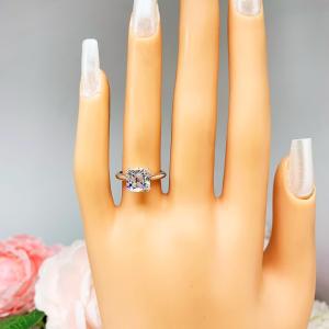 Mannequin hand with long white shimmery nails, displayed against a soft white-gray background with pink and white floral accents. The hand is wearing a sterling silver split-prong cathedral engagement ring set with a colorless old mine cut cubic zirconia.
