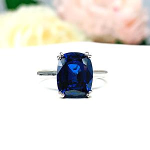 Sterling silver ring featuring a vivid elongated cushion-cut lab created blue sapphire held in a split-prong setting, with soft-focus pink and white flowers in the background.