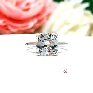 Sterling silver engagement ring with a sparkling cushion cut lab created white sapphire set in a split-prong setting set against a white surface with soft-focus pink and white flowers in the background.