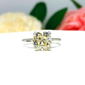 Sterling silver split prong cathedral engagement ring set with an 8mm light yellow old mine cut cubic zirconia. The ring is resting on a white surface with a background of pink and white flowers.
