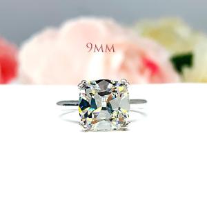 A sterling silver split-prong cathedral engagement ring set with a near colorless old mine cut cubic zirconia. The ring is set against a white background with a blurred image of pink and white flowers in the background. The text '9MM' appears in the image.