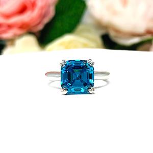 Sterling silver 8mm teal blue asscher cut cubic zirconia solitaire ring. The ring is resting on a white surface with a background of pink and white flowers.