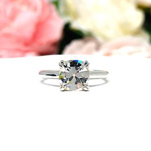 Sterling silver split prong cathedral engagement ring set with an 7.5mm near colorless old mine cut cubic zirconia. The ring is resting on a white surface with a background of pink and white flowers.