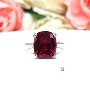 Sterling silver ring featuring a vivid elongated cushion-cut lab created ruby held in a split-prong setting, with soft-focus pink and white flowers in the background and “SJ” initials subtly visible in the corner.