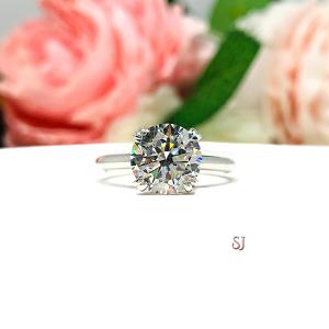 Close-up view of a near colorless (G/H/I color) round brilliant cut cubic zirconia solitaire ring in a split prong cathedral setting. The ring is made of sterling silver and features a sleek, minimalist design. The setting is simple yet elegant, allowing the center stone to take center stage. The ring is displayed against a soft, blurred background of pastel-colored flowers, creating a romantic and delicate ambiance.