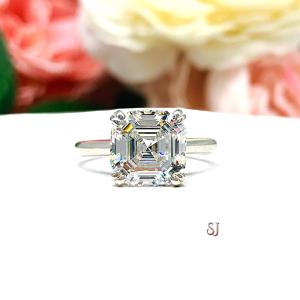 Sterling silver 9mm near colorless (G/H color) asscher cut cubic zirconia solitaire engagement ring. The ring is resting on a white surface with a background of pink and white flowers.