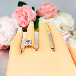 Mannequin hand with long white shimmery nails, displayed against a soft white-gray background with pink and white floral accents. The hand is wearing an elegant sterling silver cathedral engagement ring set with an 10x8mm radiant cut cubic zirconia.