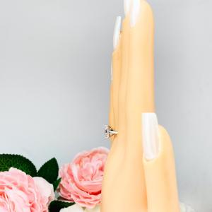 Mannequin hand with long white shimmery nails, displayed against a soft white-gray background with pink and white floral accents. The hand is wearing an elegant sterling silver cathedral engagement ring set with an 8x6mm radiant cut cubic zirconia.