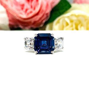 Elegant sterling silver 3-stone ring with a 10mm Asscher‑cut lab blue sapphire center and 6mm CZ side stones, displayed with soft floral backdrop.