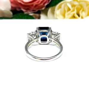 Elegant sterling silver 3-stone ring with a 10mm Asscher‑cut lab blue sapphire center and 6mm CZ side stones, displayed with soft floral backdrop.