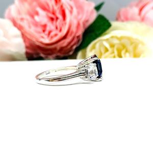 Elegant sterling silver 3-stone ring with a 10mm Asscher‑cut lab blue sapphire center and 6mm CZ side stones, displayed with soft floral backdrop.