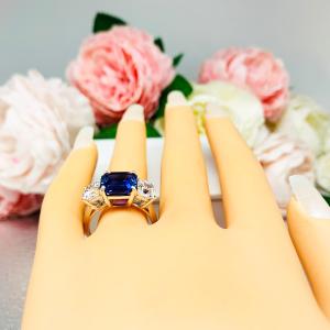 Mannequin hand with long white shimmery nails, displayed against a soft white-gray background with pink and white floral accents. The hand is wearing a sterling silver ring featuring an elegant sterling silver 3-stone ring with a 10mm Asscher‑cut lab blue sapphire center and 6mm CZ side stones.