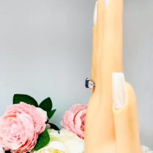 Mannequin hand with long white shimmery nails, displayed against a soft white-gray background with pink and white floral accents. The hand is wearing a sterling silver ring featuring an elegant sterling silver 3-stone ring with a 10mm Asscher‑cut lab blue sapphire center and 6mm CZ side stones.
