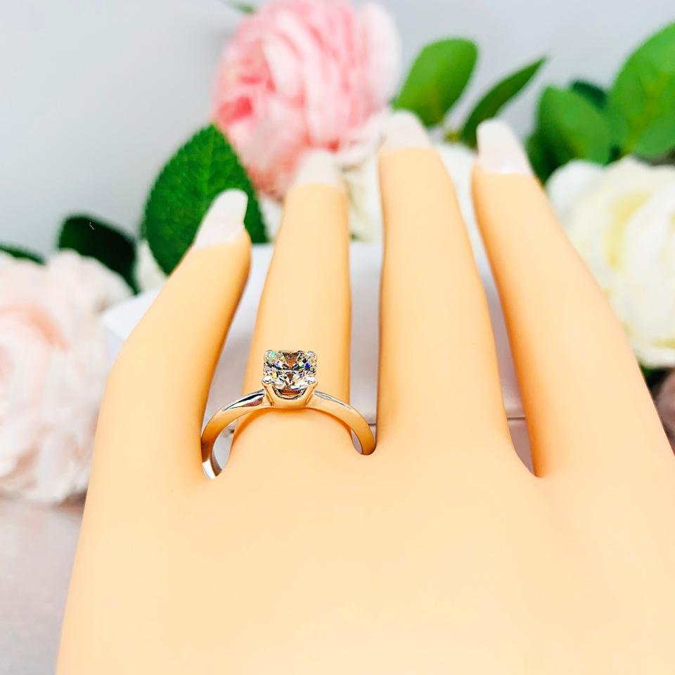 Mannequin hand with long white shimmery nails, displayed against a soft white-gray background with pink and white floral accents. The hand is wearing a sterling  silver engagement ring set with an 6mm brilliant cushion near colorless cubic zirconia.