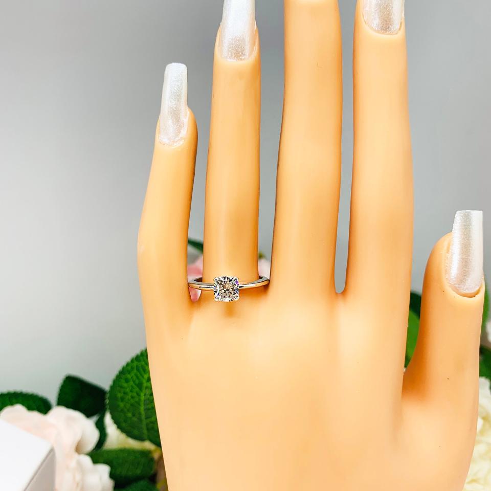 Mannequin hand with long white shimmery nails, displayed against a soft white-gray background with pink and white floral accents. The hand is wearing a sterling  silver engagement ring set with an 6mm brilliant cushion near colorless cubic zirconia.