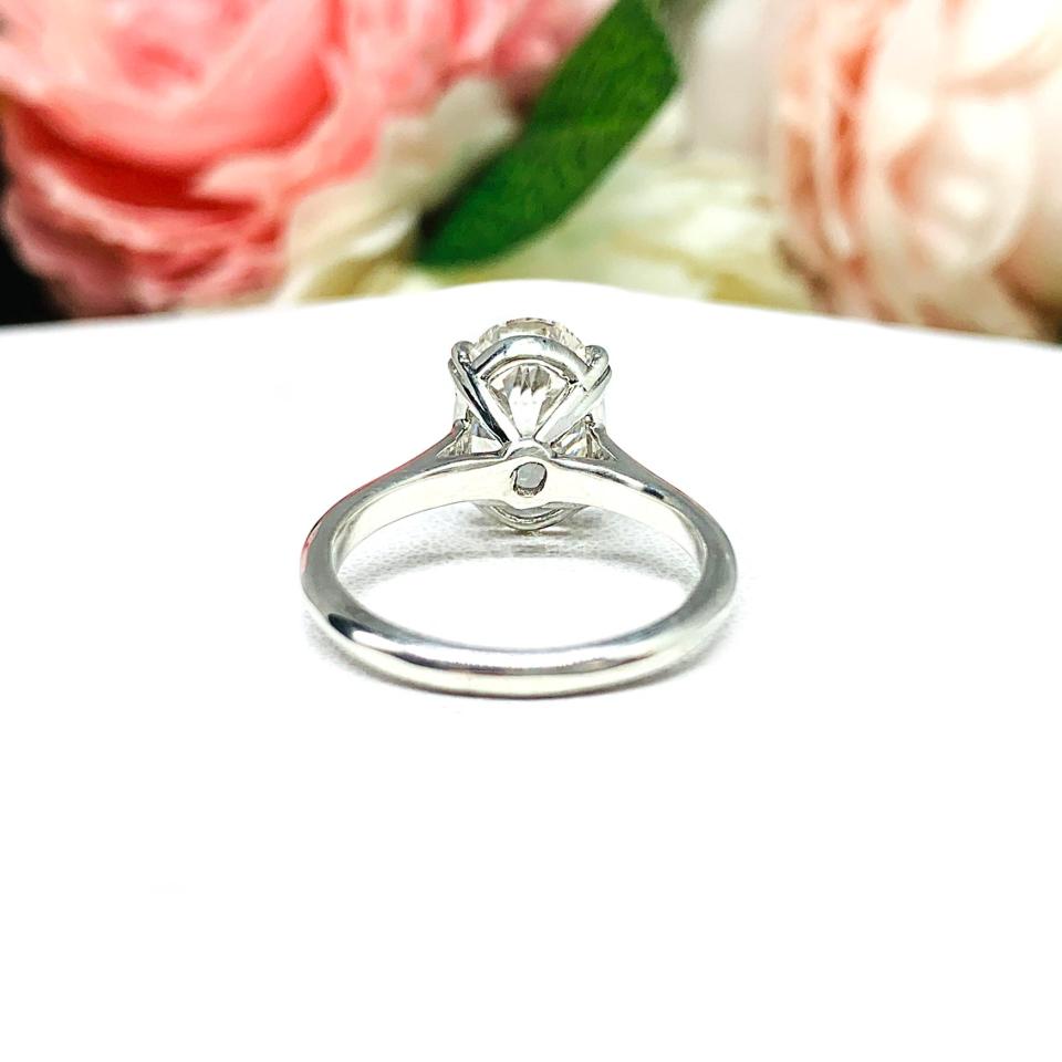 Sterling silver solitaire engagement ring set with a 12x8mm elongated oval near colorless cubic zirconia. The ring features split prongs and a cathedral shank. It is resting on a white surface with soft-focus pink and white flowers in the background.