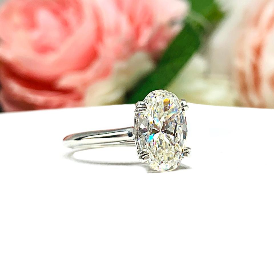 Sterling silver solitaire engagement ring set with a 12x8mm elongated oval near colorless cubic zirconia. The ring features split prongs and a cathedral shank. It is resting on a white surface with soft-focus pink and white flowers in the background.