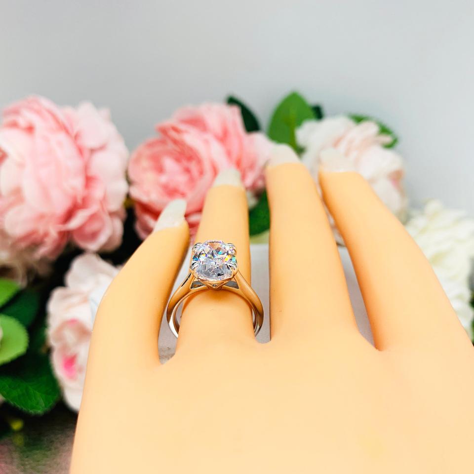 Mannequin hand with long white shimmery nails, displayed against a soft white-gray background with pink and white floral accents. The hand is wearing a sterling silver solitaire engagement ring set with a 12x8mm elongated oval cubic zirconia. The ring features split prongs and a cathedral shank.