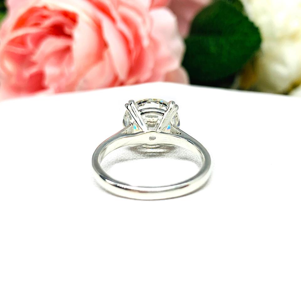 Close-up view of a near colorless (G/H/I color) round brilliant cut cubic zirconia solitaire ring in a split prong cathedral setting. The ring is made of sterling silver and features a sleek, minimalist design. The setting is simple yet elegant, allowing the center stone to take center stage. The ring is displayed against a soft, blurred background of pastel-colored flowers, creating a romantic and delicate ambiance.