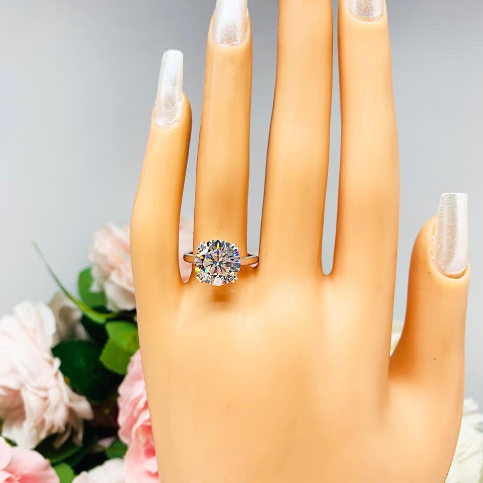 Mannequin hand with long white shimmery nails, displayed against a soft white-gray background with pink and white floral accents. The hand is wearing a near colorless (G/H/I color) round brilliant cut cubic zirconia solitaire ring in a split prong cathedral setting. The ring is made of sterling silver and features a sleek, minimalist design. The setting is simple yet elegant, allowing the center stone to take center stage.