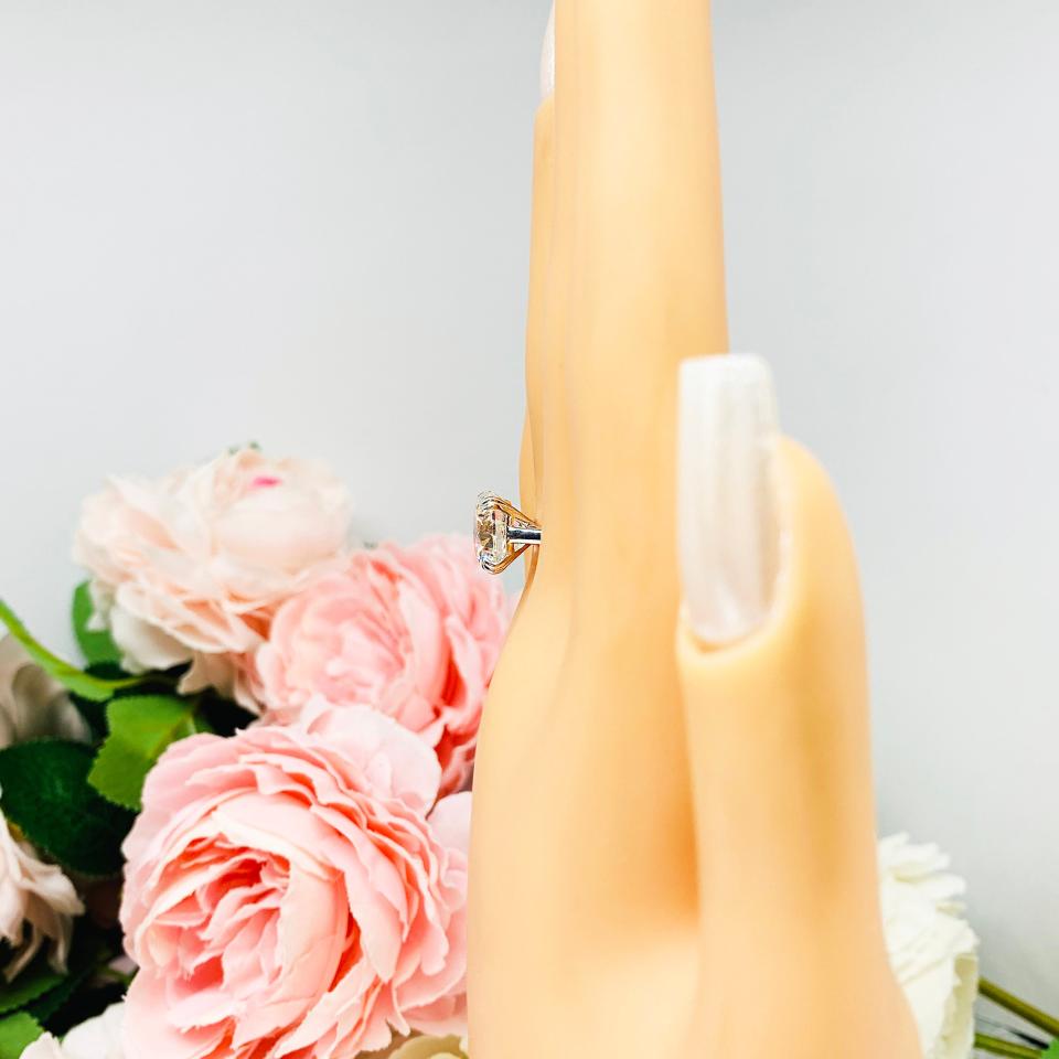 Mannequin hand with long white shimmery nails, displayed against a soft white-gray background with pink and white floral accents. The hand is wearing a near colorless (G/H/I color) round brilliant cut cubic zirconia solitaire ring in a split prong cathedral setting. The ring is made of sterling silver and features a sleek, minimalist design. The setting is simple yet elegant, allowing the center stone to take center stage.