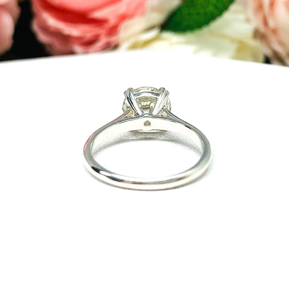 Close-up view of a near colorless (G/H/I color) round brilliant cut cubic zirconia solitaire ring in a split prong cathedral setting. The ring is made of sterling silver and features a sleek, minimalist design. The setting is simple yet elegant, allowing the center stone to take center stage. The ring is displayed against a soft, blurred background of pastel-colored flowers, creating a romantic and delicate ambiance.