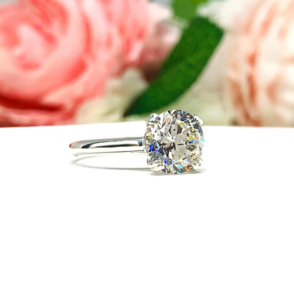 Close-up view of a near colorless (G/H/I color) round brilliant cut cubic zirconia solitaire ring in a split prong cathedral setting. The ring is made of sterling silver and features a sleek, minimalist design. The setting is simple yet elegant, allowing the center stone to take center stage. The ring is displayed against a soft, blurred background of pastel-colored flowers, creating a romantic and delicate ambiance.