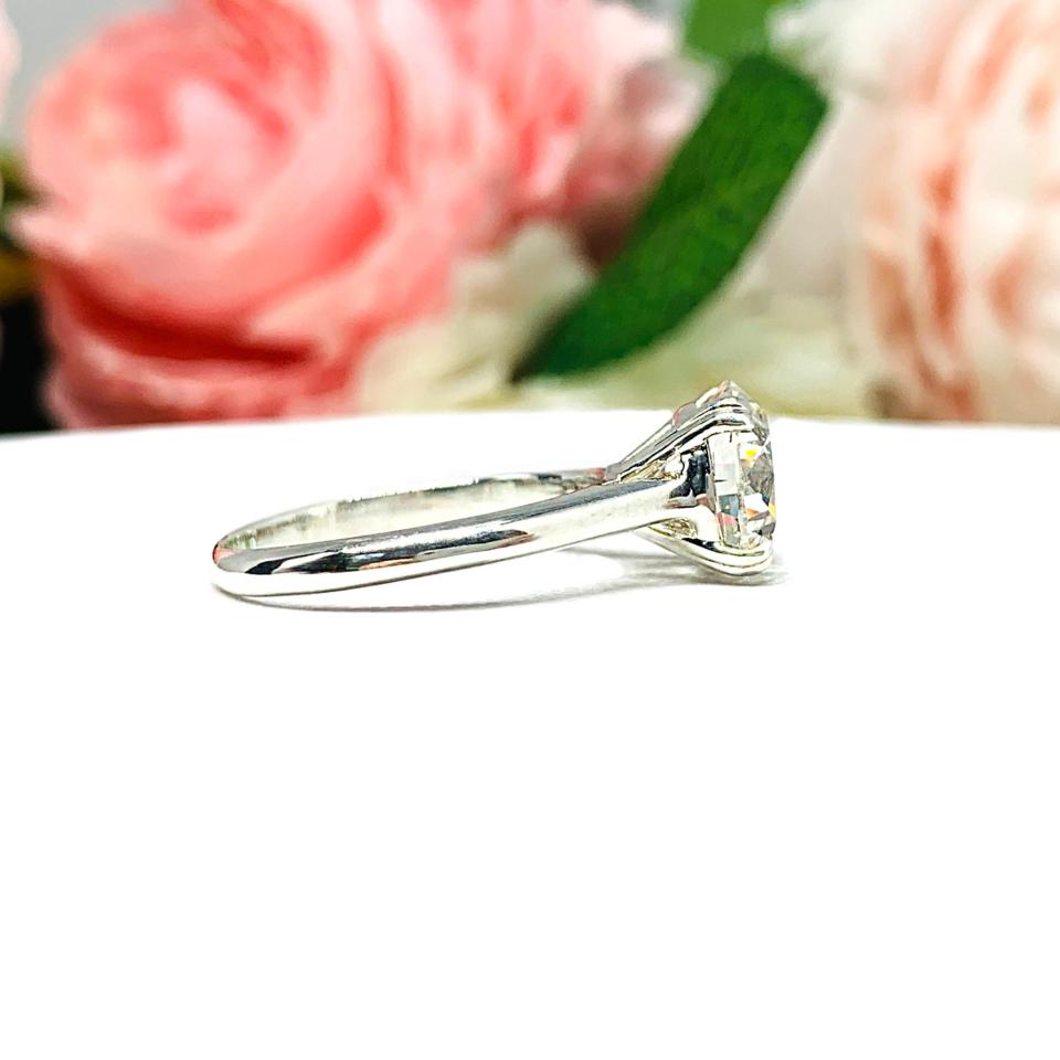 Close-up view of a near colorless (G/H/I color) round brilliant cut cubic zirconia solitaire ring in a split prong cathedral setting. The ring is made of sterling silver and features a sleek, minimalist design. The setting is simple yet elegant, allowing the center stone to take center stage. The ring is displayed against a soft, blurred background of pastel-colored flowers, creating a romantic and delicate ambiance.