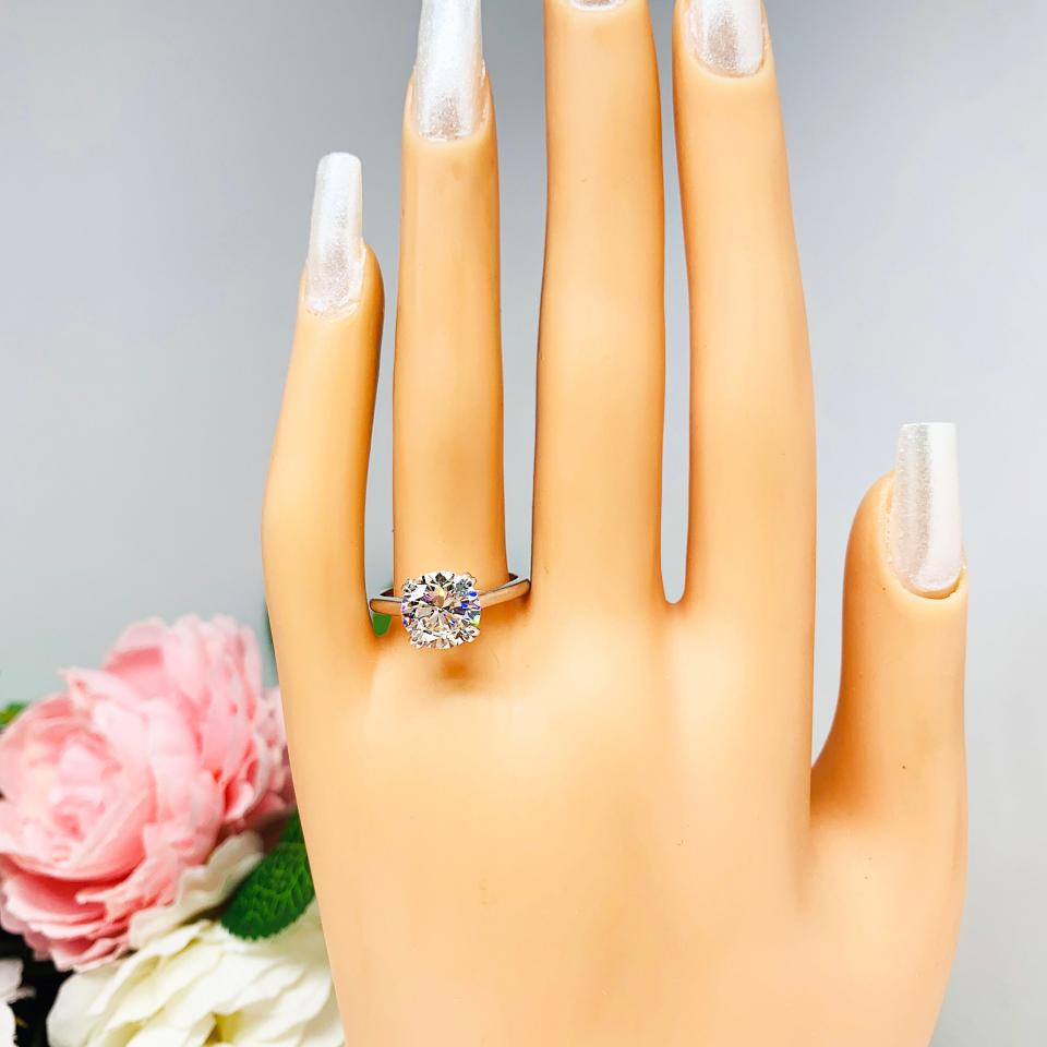 Mannequin hand with long white shimmery nails, displayed against a soft white-gray background with pink and white floral accents. The hand is wearing a near colorless (G/H/I color) round brilliant cut cubic zirconia solitaire ring in a split prong cathedral setting. The ring is made of sterling silver and features a sleek, minimalist design. The setting is simple yet elegant, allowing the center stone to take center stage.