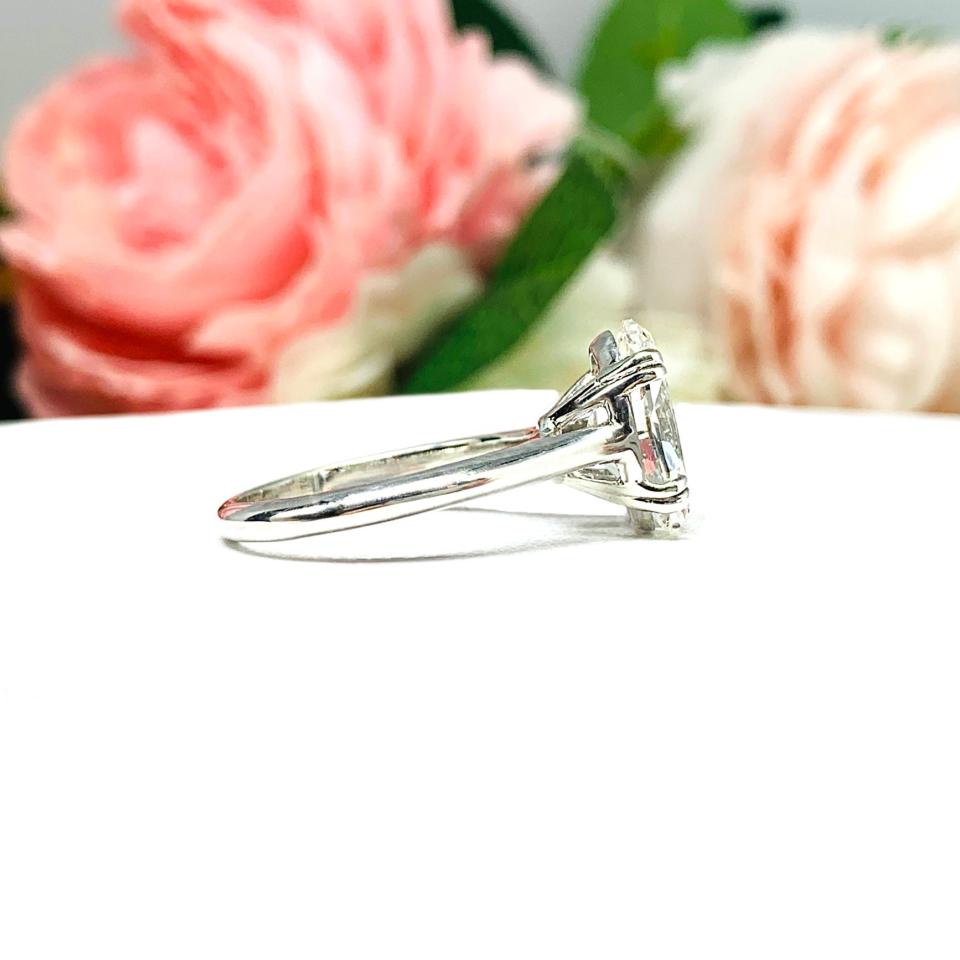 Sterling silver solitaire engagement ring set with a 12x8mm elongated oval near colorless cubic zirconia. The ring features split prongs and a cathedral shank. It is resting on a white surface with soft-focus pink and white flowers in the background.