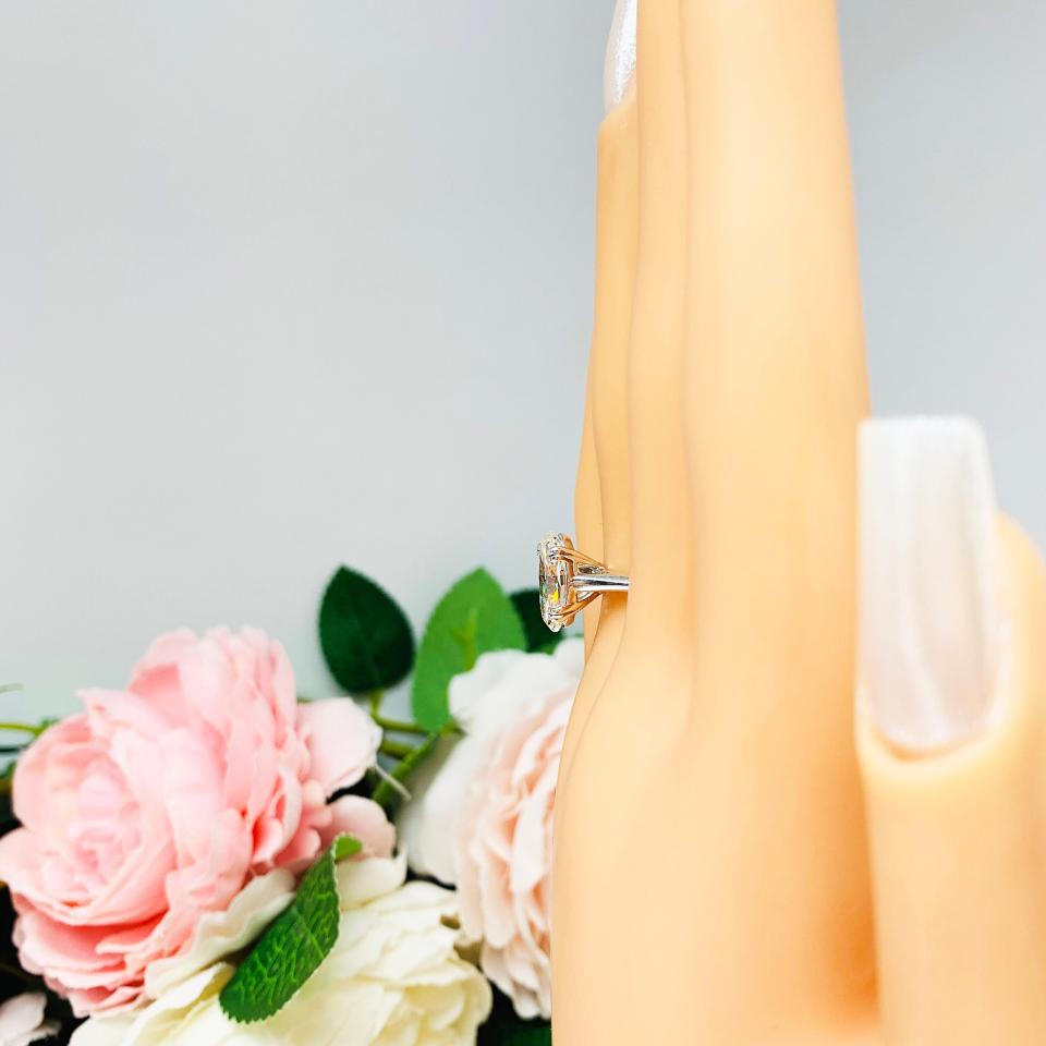 Mannequin hand with long white shimmery nails, displayed against a soft white-gray background with pink and white floral accents. The hand is wearing a sterling silver solitaire engagement ring set with a 12x8mm elongated oval near colorless cubic zirconia. The ring features split prongs and a cathedral shank.