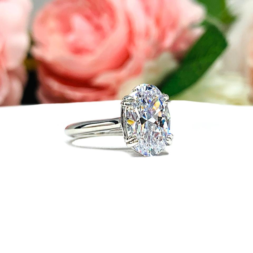 Sterling silver solitaire engagement ring set with a 12x8mm elongated oval cubic zirconia. The ring features split prongs and a cathedral shank. It is resting on a white surface with soft-focus pink and white flowers in the background.