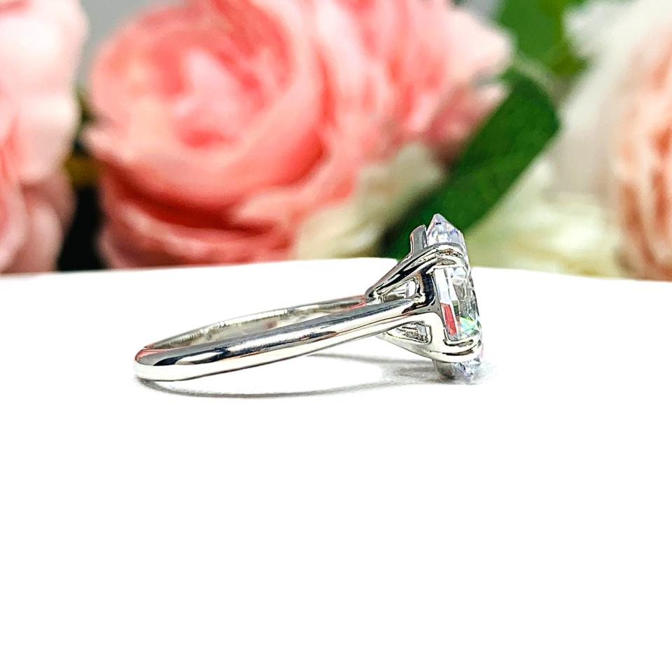 Sterling silver solitaire engagement ring set with a 12x8mm elongated oval cubic zirconia. The ring features split prongs and a cathedral shank. It is resting on a white surface with soft-focus pink and white flowers in the background.