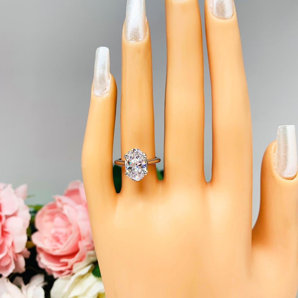 Mannequin hand with long white shimmery nails, displayed against a soft white-gray background with pink and white floral accents. The hand is wearing a sterling silver solitaire engagement ring set with a 12x8mm elongated oval cubic zirconia. The ring features split prongs and a cathedral shank.