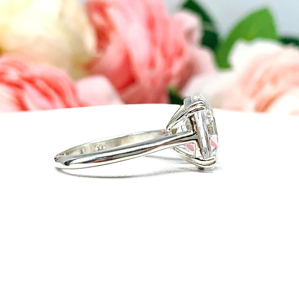 Sterling silver engagement ring featuring a sparkling elongated cushion-cut lab created white sapphire held in a split-prong setting, with soft-focus pink and white flowers in the background.