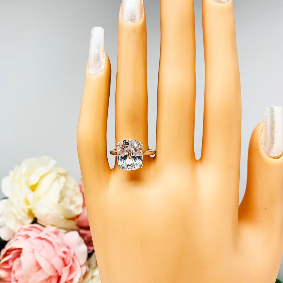 Mannequin hand with long white shimmery nails, displayed against a soft white-gray background with pink and white floral accents. The hand is wearing a sterling silver engagement ring featuring a sparkling elongated cushion-cut lab created white sapphire held in a split-prong setting.