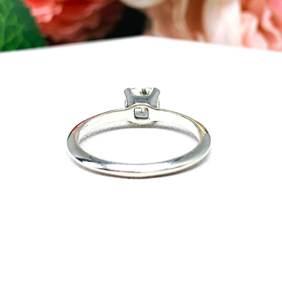 Sterling silver engagement ring set with an 6mm brilliant cushion near colorless cubic zirconia. The ring is resting on a white surface with a background of soft-focus pink and white flowers.