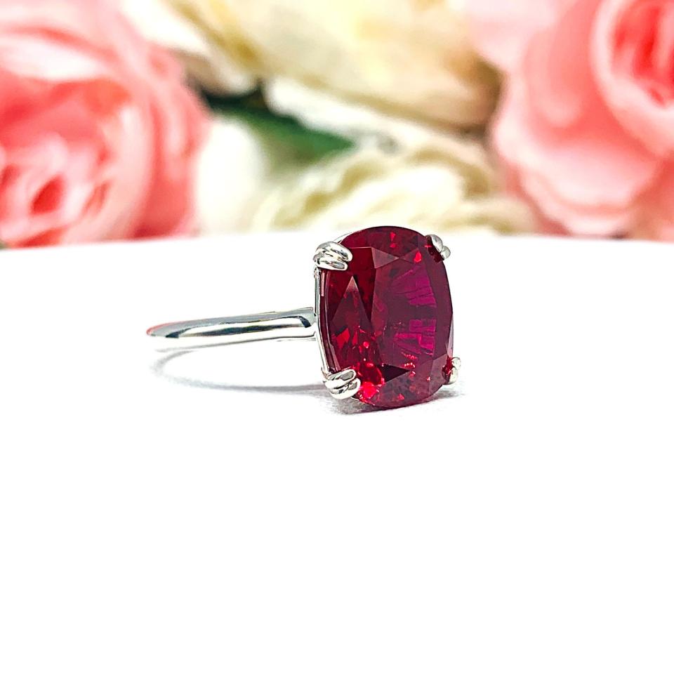 Sterling silver ring featuring a vivid elongated cushion-cut lab created ruby held in a split-prong setting, with soft-focus pink and white flowers in the background and “SJ” initials subtly visible in the corner.